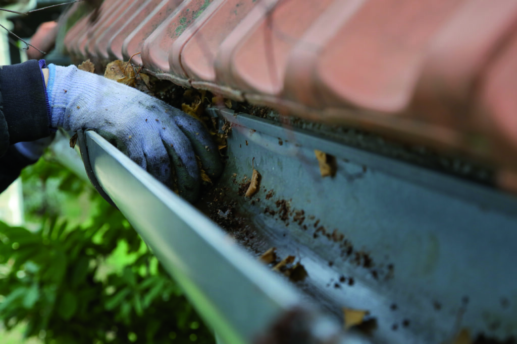 A person wearing work gloves is cleaning leaves and debris from a rain gutter along a tiled roof.